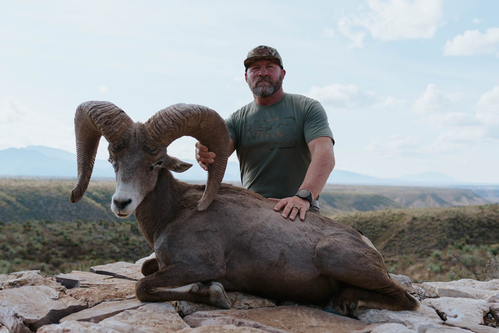 A hunter kneeling behind a full-curl desert bighorn ram on white rock, a Coahuila canyon falling away behind