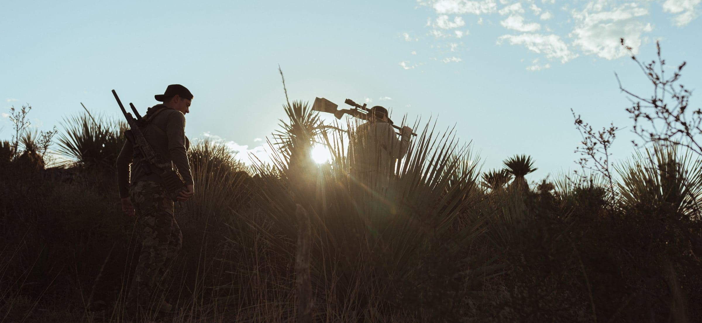 Two hunters cresting a yucca ridge at sunset, one on a spotting scope, the sun starring behind them — glassing the Mexican desert