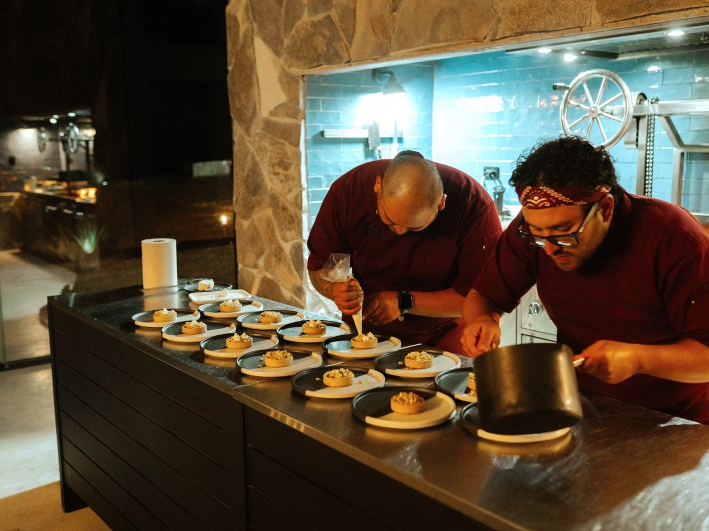 Two chefs in burgundy plating a long row of desserts at the stone open kitchen