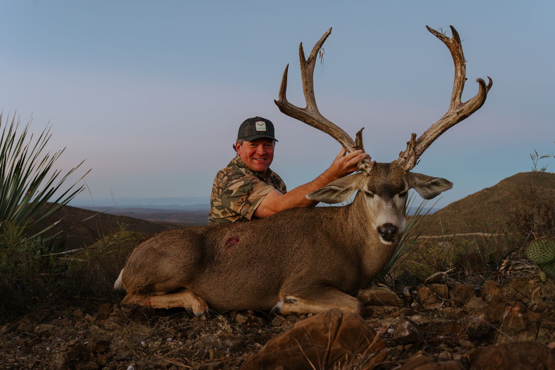 A hunter beside a heavy desert mule deer buck on a Coahuila summit at lavender dusk, layered mountains behind